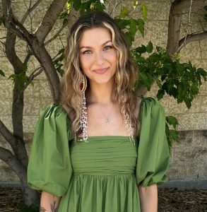 Image of woman with longer brown hair, wearing a green top, facing the camera. Background is trees in front of a brick wall. 